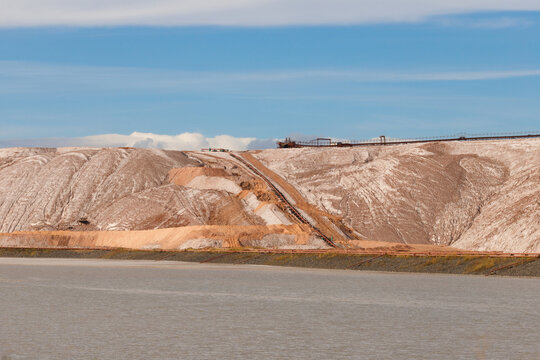 Massive textured brown and white salt slag heaps of a potash mining quarry with a long conveyor belt system stretching along the ridge under a blue sky horizontal landscape view