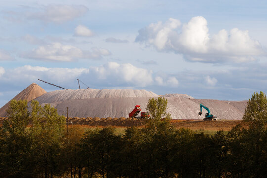 Red dump truck and blue excavator working on a soil embankment with massive white salt slag heaps on the background under a cloudy sky horizontal landscape view