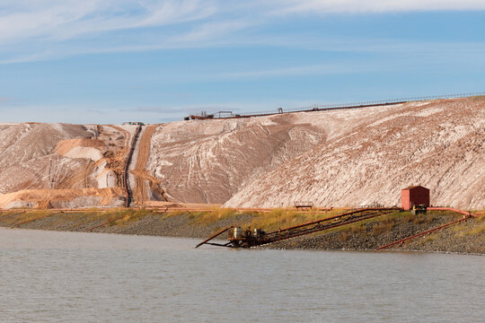 Massive brown and white salt slag heaps of a potash mining quarry with a long conveyor belt system rising behind a technical water reservoir pond under a blue sky horizontal view