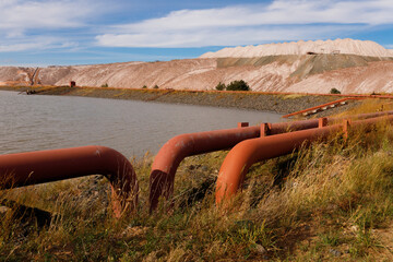 Multiple industrial rusty iron pipelines with curved u shaped bends on the grass shore of a technical reservoir pond near massive brown salt slag heaps under a cloudy blue sky wide horizontal view © Tina