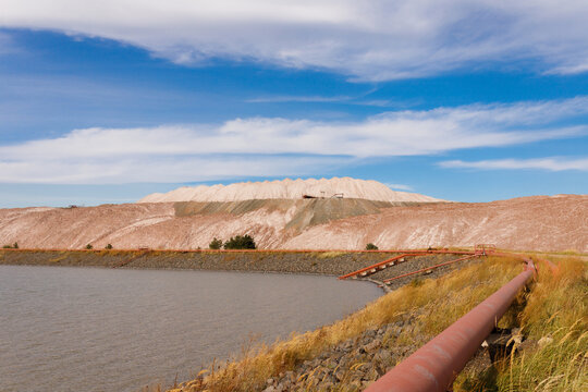 Wide landscape view of a technical water reservoir pond with an industrial rusty iron pipeline on the grass shore and massive brown and grey salt slag heaps under a cloudy blue sky