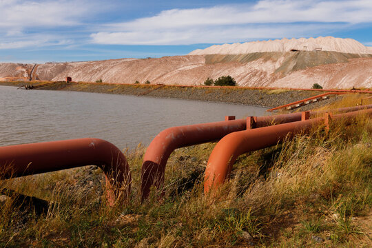 Multiple industrial rusty iron pipelines with curved u shaped bends on the grass shore of a technical reservoir pond near massive brown salt slag heaps under a cloudy blue sky wide horizontal view