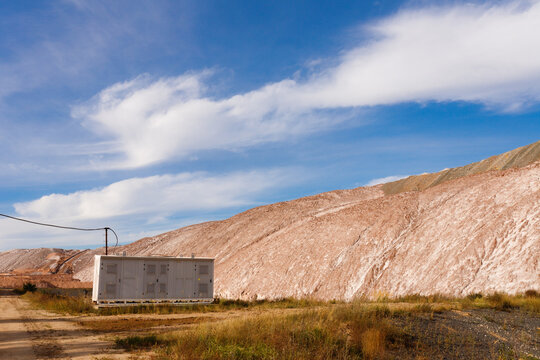 Metal package transformer substation kiosk next to a rural dirt road with massive industrial brown and white potash salt slag heaps on the background under a blue cloudy sky horizontal view
