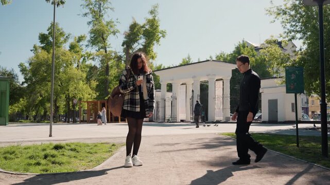 Young woman looks down at her smartphone while standing outdoors in daylight with trees visible behind her. Another person is seen walking away from camera and later man approaches to engage