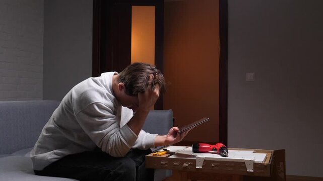 Young man on sofa covers his face in frustration while consulting a tablet amid unassembled furniture pieces, tools and boxes during a tricky diy assembly project