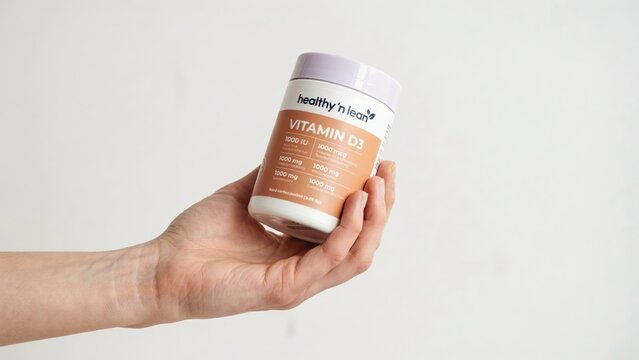 A hand holding a white plastic bottle of vitamin d3 supplement with a peach colored label against a plain light gray studio background, wall surface