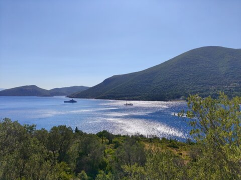 Panoramic coastal view of Ithaca island, Greece, with shimmering blue sea, green hills and sailing boats under clear summer sky