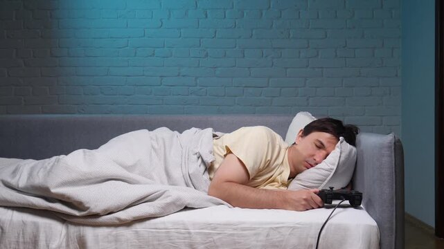 Exhausted man sleeping on a couch, holding a video game controller in his hand, illustrating themes of gaming addiction, burnout, and excessive screen time late at night