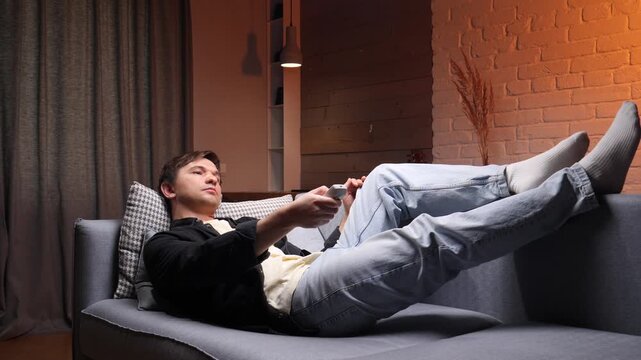 Man reclining on a comfortable sofa in his living room with feet up, holding a remote control and watching tv, enjoying a relaxed evening at home after work