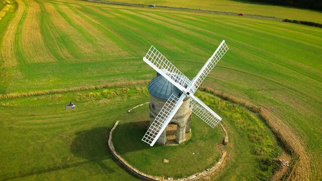 Drone view of historic stone windmill with white sails among farmland. Traditional rural mill from above inside circular stone wall. Architectural heritage monument amid green cultivated countryside