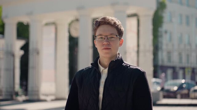 Young adult male wearing glasses stands alone in urban public square looking down and to side with contemplative expression. Man is dressed casually but neatly black quilted jacket