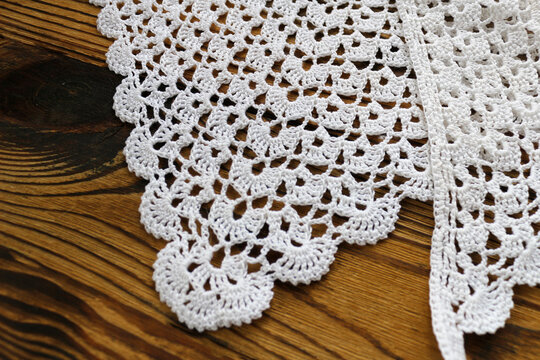 Elegant white handmade crochet lace with delicate scalloped border pattern laid on a rustic dark wooden table. Perfect background texture for craft, vintage, needlework, and home decor projects.