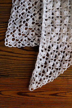 Elegant white handmade crochet lace with delicate scalloped border pattern laid on a rustic dark wooden table. Perfect background texture for craft, vintage, needlework, and home decor projects.