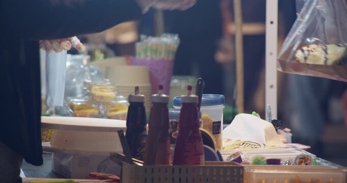 A gloved street food vendor packages a sweet dessert in a plastic bag beside condiment bottles at a Thai night market stall.