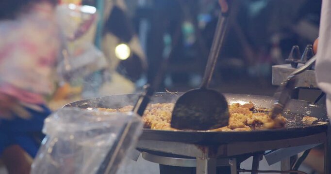 A street food vendor stir-fries oyster omelette with egg batter on a sizzling flat pan at a Thai night market stall.