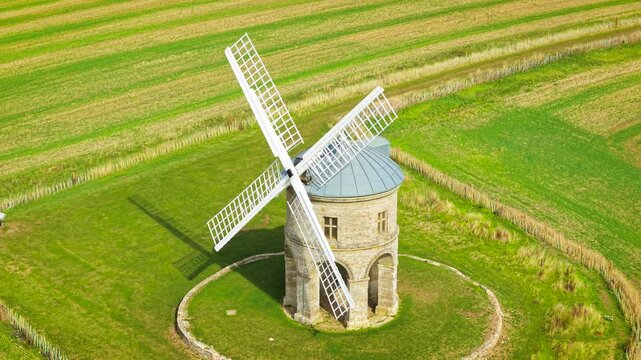 Drone view of traditional stone windmill for flour milling. Historic wheat and grain mill with arched base, metal roof and lattice sails. Rural milling structure within circular lawn beside cultivated