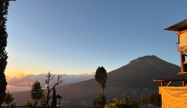 Majestic volcanic mountain rises above glowing clouds at sunrise, while warm golden light illuminates the rugged slopes, trees, and rustic buildings surrounding the scenic viewpoint.
