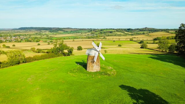 Old wheat windmill for traditional flour milling. Small historic stone grain mill with white lattice sails on open green hillside. Rural agricultural landmark among patchwork fields and rolling