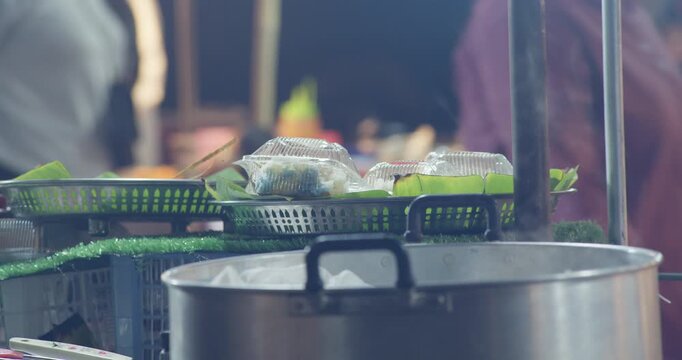A street food vendor reaches past a steaming pot toward banana leaf-wrapped desserts packed in plastic containers at a night market.