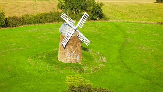 Historic wheat windmill and traditional flour mill. Old stone grain milling tower with white lattice sails and weathered masonry. Rural agricultural structure on green pasture beside hedgerows and