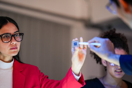 An engineer raises the die on tweezers for a closer look with a colleague watching from just behind her during a focused late-night working session with the rest of the team nearby