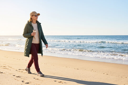 Woman in warm parka jacket and cap walking along sandy ocean beach carrying coffee cup