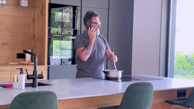 Mature man with glasses and a beard simultaneously talking on the phone and stirring food in a pot while standing in a modern, well-equipped kitchen, showcasing multitasking at home