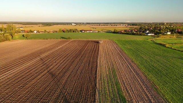 Aerial view of tractor cultivating field with fresh furrows next to green crop field at golden hour sunset