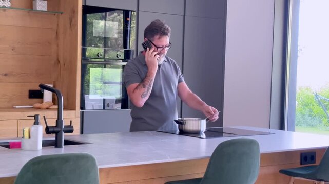 Multitasking man with a beard and glasses talking on the phone while cooking a meal in his modern kitchen, stirring the contents of a pot on an induction stove with a wooden spoon