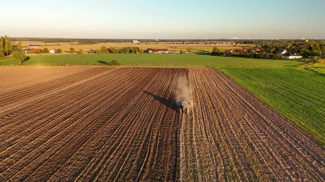 Aerial view of tractor with harrow cultivating soil and raising dust next to green field at sunset