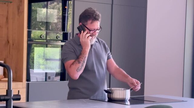 Middle-aged man with glasses and a beard talking on the phone while cooking a meal in his modern kitchen, multitasking by stirring a pot on the induction stove during his conversation