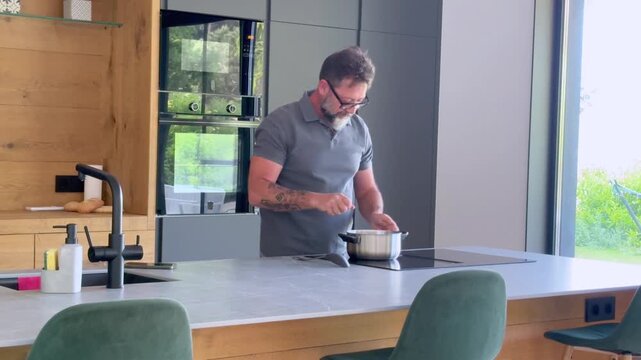 Confident middle-aged man with a beard and glasses preparing food in a saucepan on a modern induction cooktop, enjoying a quiet moment while cooking a healthy meal at home
