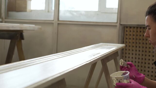 Craftswoman painter painting vintage wooden door panel with brush in paint booth in carpentry workshop, creative working process of furniture restoration, woman painting renovated wooden door.