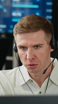 Vertical video of Caucasian young adult software specialist wearing headset talking while monitoring data on computer screen. Footage suitable for customer service and tech support advertising