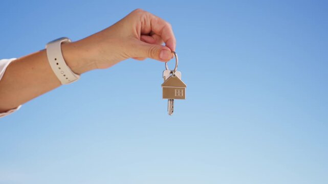 Woman's hand holding a new house key with a keychain against a clear blue sky, symbolizing real estate investment, property ownership, and buying a home for the family