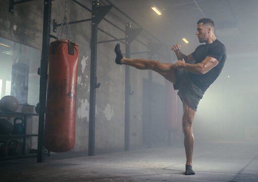 Powerful muscular male athlete kicking a heavy punching bag with flying dust in a dark smoky gym, or intense martial arts combat training for active fitness lifestyle.