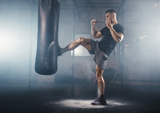 Powerful muscular male athlete kicking a heavy punching bag with flying dust in a dark smoky gym, or intense martial arts combat training for active fitness lifestyle.