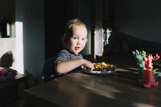 Cute toddler boy eats porridge with mango while playing with colorful toy dinosaurs at a home dining table. Authentic childhood lifestyle concept showing playful everyday mealtime moments.