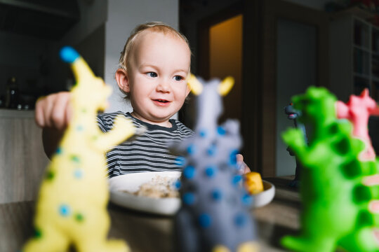 Cute toddler boy eats porridge with mango while playing with colorful toy dinosaurs at a home dining table. Authentic childhood lifestyle concept showing playful everyday mealtime moments.