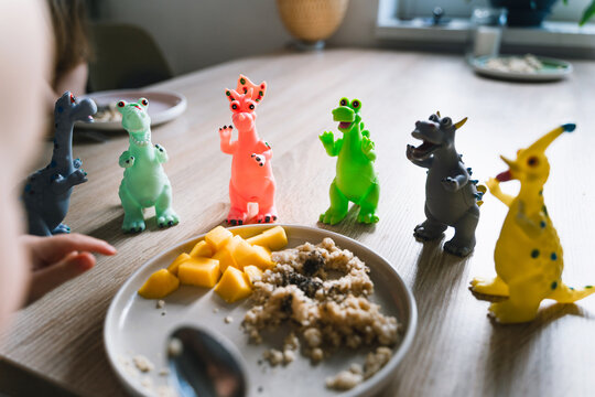 Cute toddler boy eats porridge with mango while playing with colorful toy dinosaurs at a home dining table. Authentic childhood lifestyle concept showing playful everyday mealtime moments.