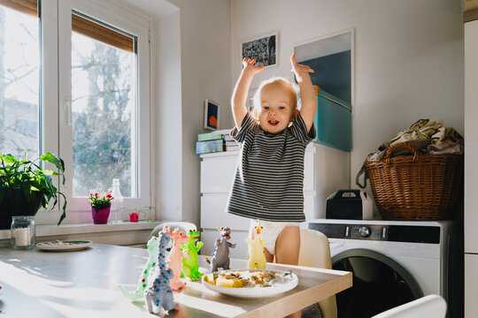 Cute toddler boy eats porridge with mango while playing with colorful toy dinosaurs at a home dining table. Authentic childhood lifestyle concept showing playful everyday mealtime moments.