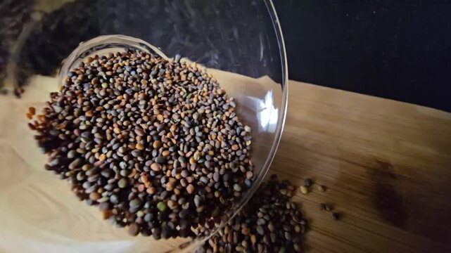 Close-up macro shot of organic arugula and rocket seeds spilling from a transparent glass bowl onto a wooden surface, creating an appealing agricultural and culinary visual.