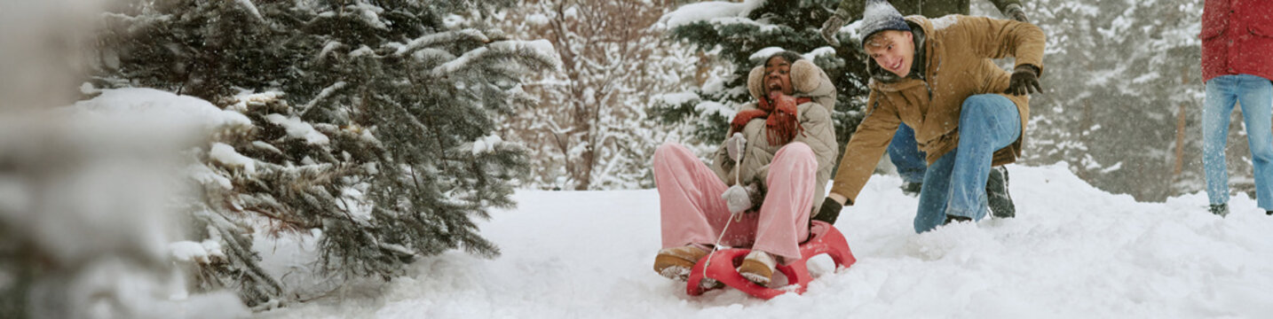Header of multiethnic young adult friends sledding outdoors on snowy slope in winter forest. Image suitable for winter vacation marketing, seasonal recreation, youth lifestyle campaigns