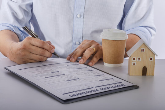 Focused businessman signs housing mortgage finance process document with wooden house model on office desk