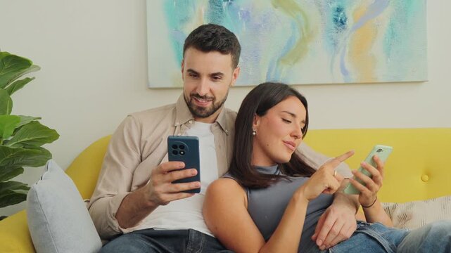 A cozy domestic moment shows a man and woman on a yellow cushioned sofa, each absorbed in their screens. Soft daylight and calm decor frame their relaxed posture and light interaction across the room.