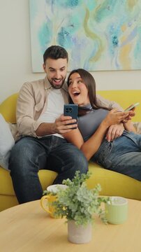 A cheerful couple lounges on a bright yellow sofa, looking at their smartphones with relaxed smiles and amused expressions. Natural light fills a cozy living room while decorative plants sit on a