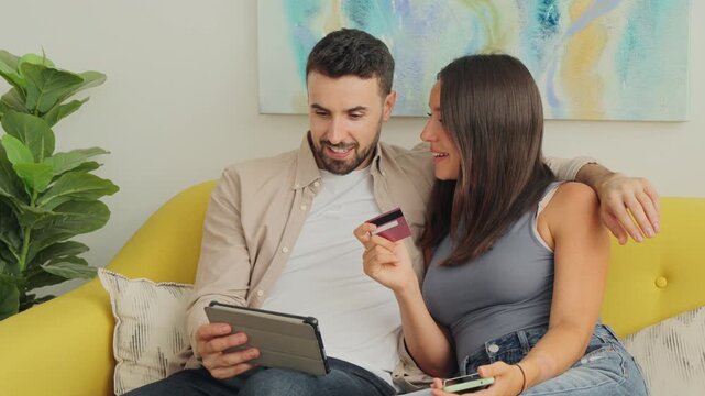 A cheerful couple sits on a yellow sofa using a tablet to shop online while one holds a credit card. Bright sunlit room and soft cushions frame a relaxed home shopping moment for media and lifestyle