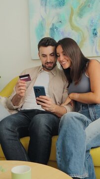 Two adults sit together on a yellow sofa in a sunlit room, using a tablet for online shopping as one holds a card, smiling and sharing a moment of digital purchase