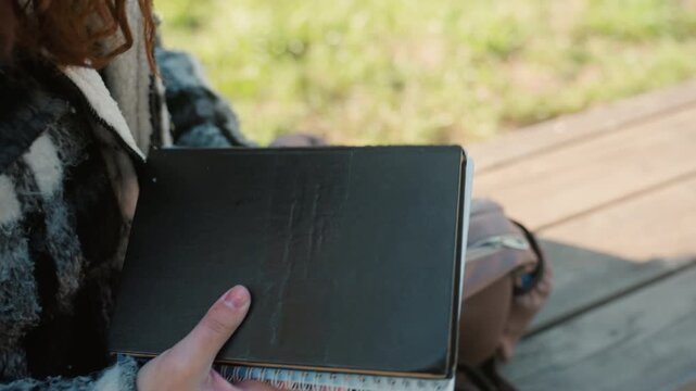 Close up shows woman with red hair wearing checkered jacket hands placing thick black notebook into pink and brown backpack on wooden bench outdoors. Notebook has lined pages visible