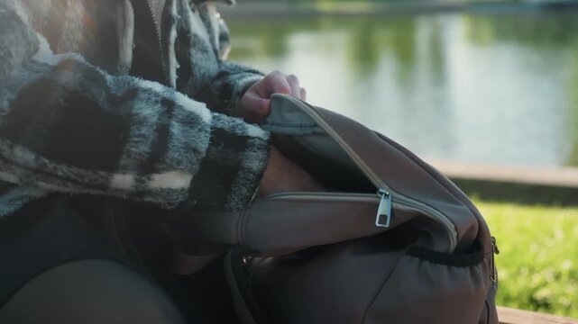 Person in checkered jacket sits by lake reaching into their brown backpack to retrieve various personal belongings. scene is set outdoors on sunny day near calm waters with green grass